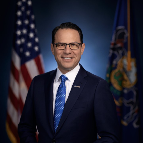 An official headshot of Governor Josh Shapiro. He is sitting in front of an American flag and the flag of the Commonwealth of Pennsylvania. 