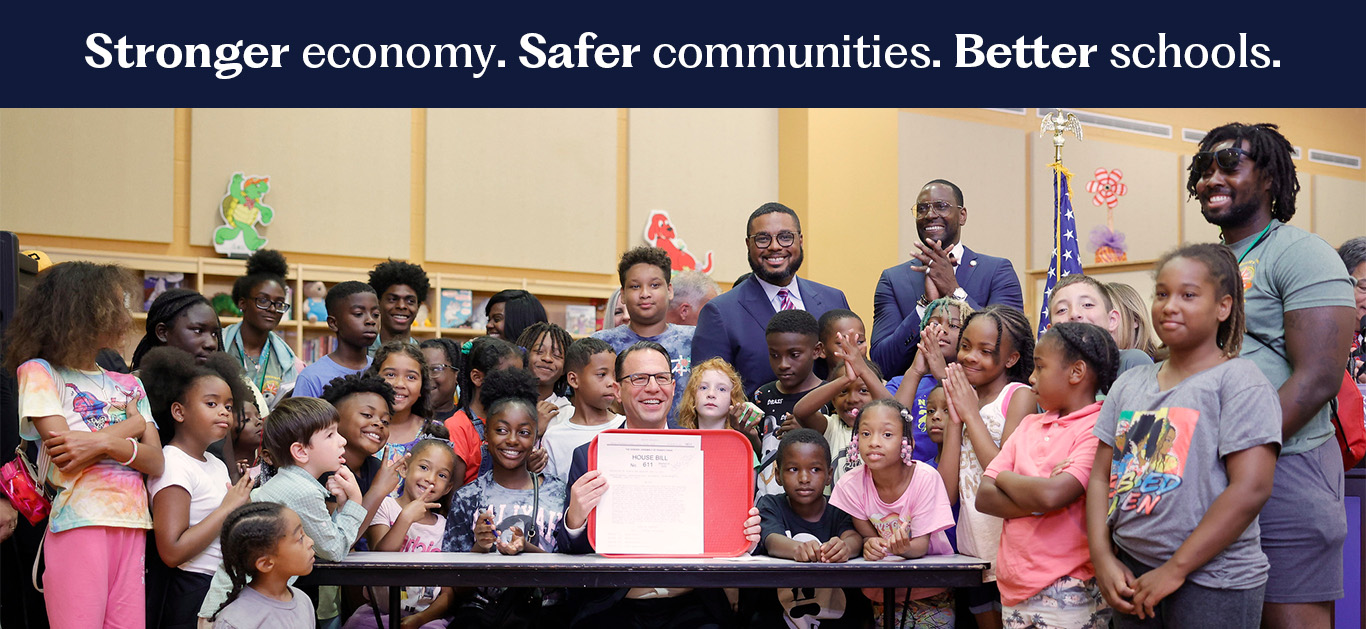 A photograph of Governor Josh Shapiro and Lt. Governor Austin Davis with elementary school children. Governor Shapiro is holding House Bill 611. This House Bill gives free school breakfast to all Pennsylvania public school students. There are words at the top of the photograph that read "stronger economy, safer communities, and better schools." 