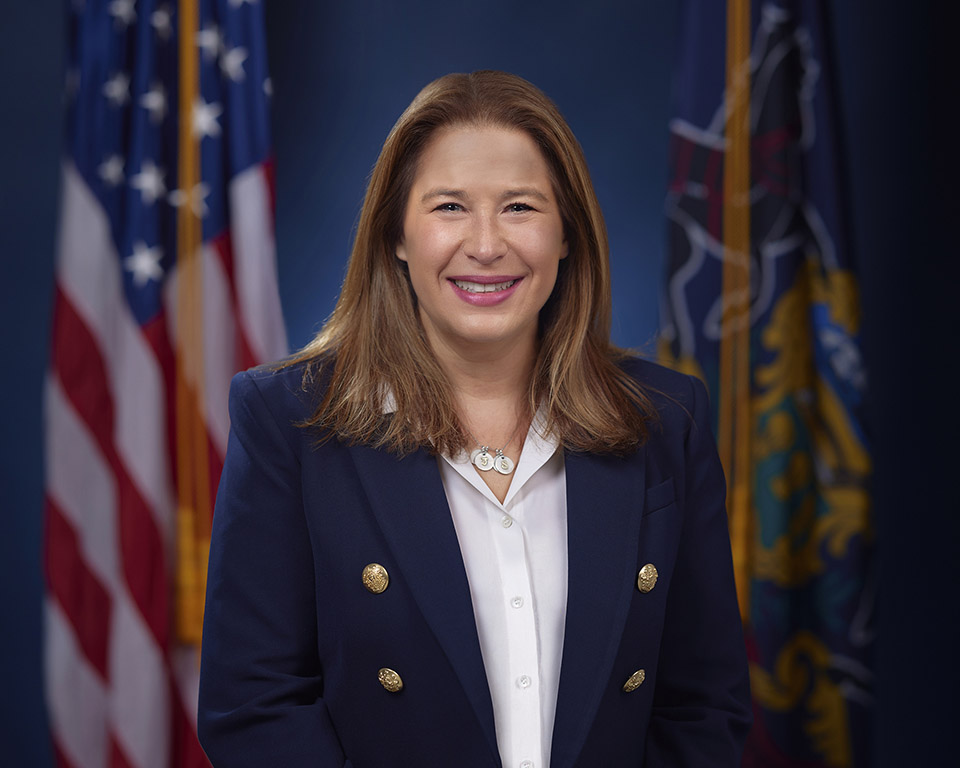 An official headshot of the First Lady Lori Shapiro. The First Lady is sitting in front of the American flag and the flag of the Commonwealth of Pennsylvania. 