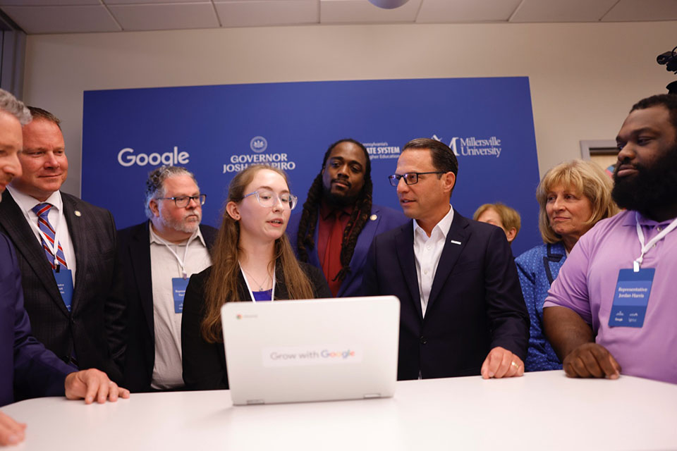 A photograph of Governor Josh Shapiro standing at a laptop with a student and a group of people in Millersville Pennsylvania. 