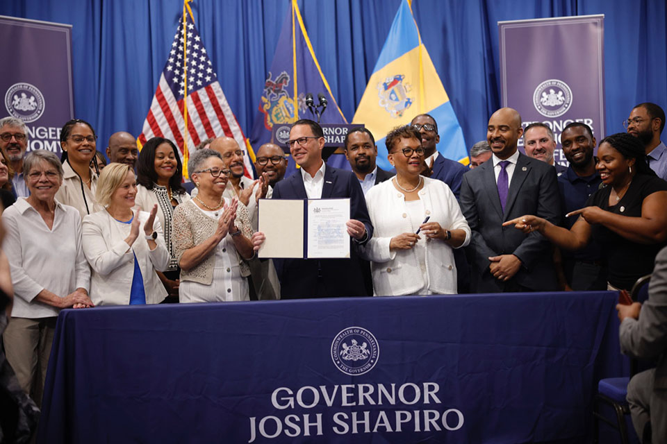 A photograph of Governor Shapiro holding up a signed bill surrounded by small diverse business owners. 