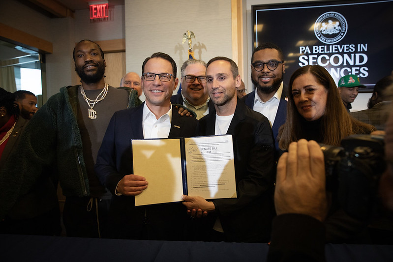 A photograph of Governor Shapiro holding up a signed bill in front of Lt. Governor Austin Davis, and Meek Mill. 